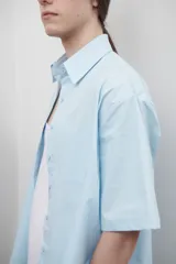 Camisa celeste de popelina de algodón, corte amplio, con cuello clásico, manga corta y bolsillo frontal. Presenta canesú con pliegue central en la espalda.