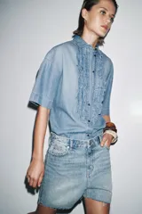 Camisa de denim celeste con cuello solapa y manga corta. Presenta detalle de volantes en el frente y bajo asimétrico con aberturas laterales.