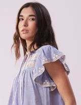 Blusa blanca de algodón con cuello a la base y escote en V con lazo de ajuste. Tiene mangas cortas con terminación amplia y bordados en delantera, mangas y espalda.