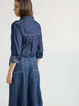 Camisa celeste de denim suave, corte relajado, con cuello clásico, cierre frontal con botones marrones, manga larga con puños abotonados, dos bolsillos de parche en el pecho y ruedo redondeado.