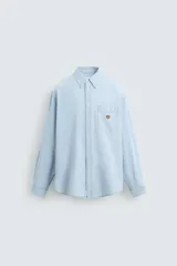 Camisa celeste de denim ligero de algodón, corte relaxed fit, cuello abotonado, manga larga con puño abotonado, bolsillo de parche en el pecho con bordado.
