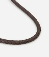 Bolso de playa tipo canasta, tejido en fibra de papel color marrón oscuro, con forma redondeada y abertura superior curva. Cuenta con asas trenzadas de material sintético marrón oscuro y detalles de flecos colgantes a los lados.