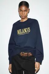 Sudadera de tejido de punto color azul marino, con cuello redondo y mangas raglán. Presenta un estampado frontal en amarillo con el texto "MILANO 75" y una dirección debajo.