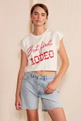 Remera corta tipo crop de algodón orgánico color blanco, con mangas cortas de terminación rústica y estampa frontal en color rojo con la frase 'But first, RODEO'.