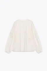 Camisa color crudo confeccionada en mezcla de algodón y lino, con cuello redondo y escote pico ajustable con lazadas. Tiene manga larga abullonada y detalle de bordados perforados a tono.
