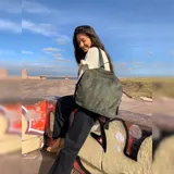 Mochila color verde militar, agamuzada con detalle tramado al frente. Apertura superior con cierre metálico. Incluye división al medio y bolsillos internos. Correa corta de mano y correas para colgar en la espalda regulables.