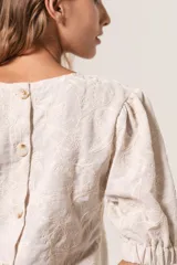 Blusa color crema con bordado de hojas, escote redondo, mangas 3/4 abullonadas con elástico en los puños y cierre con botones en la espalda.