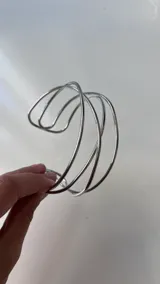 Brazalete abierto de metal plateado, estilo cuff, compuesto por múltiples alambres delgados que se cruzan y entrelazan de forma abstracta.