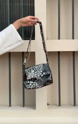 Cartera bandolera negra de ecocuero con textura arrugada, tachas plateadas, cadena decorativa y aro metálico.