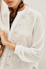 Camisa blanca de manga larga con cuello mao y escote en V. Presenta bordados florales en blanco y dorado en el frente.