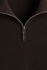 Buzo de punto color marrón oscuro (casi negro), de manga larga, con cuello alto y medio cierre metálico frontal. Presenta un tejido acanalado en el cuello y un tejido liso en el cuerpo.