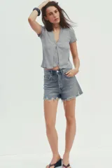 Camiseta color beige de tejido de punto, con cuello halter y escote pico. Tiene cierre frontal con botones y mangas cortas.