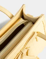 Cartera mini city color amarillo claro, de cuerina trenzada, con doble asa corta de mano, correa regulable desmontable, cierre superior y moño decorativo.