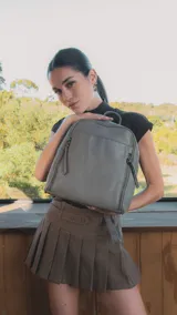 Mochila gris con detalle de costuras en forma de rombos en el frente. Tiene un bolsillo frontal con broche magnético, asa de mano y correas regulables.