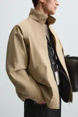 Parka ligera color beige, de corte relaxed fit, confeccionada en tejido técnico repelente al agua. Tiene cuello alto, mangas largas con puños ajustables con botón a presión, bolsillos de solapa en la cadera y cierre frontal con cremallera oculta por solapa con botones a presión.