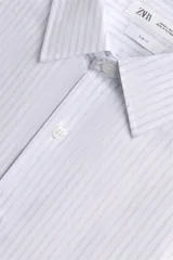 Camisa slim fit blanca con rayas verticales finas en color gris, confeccionada en tejido de algodón. Presenta cuello italiano y manga larga con puño abotonado. Cierre frontal con botonadura.