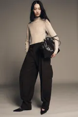 Pantalón cargo de algodón color marrón oscuro, tiro medio, con pliegues frontales, bolsillos delanteros ocultos y bolsillos de plastrón con solapa y botones a presión en las piernas. El bajo del pantalón tiene una trabilla con botón y se muestra doblado hacia afuera.