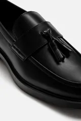 Mocasín de vestir color marrón oscuro, con detalle de borlas en la parte delantera, horma redonda y suela con ligero tacón.
