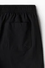 Short de baño negro de sirsaca con aberturas laterales cortas, cintura elástica con cordón ajustable, bolsillos laterales y bolsillo trasero con cierre autoadherente. Tiene suspensor interior de malla.