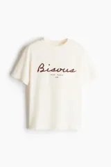Remera color crema de corte holgado en punto de algodón. Modelo con cuello redondo y estampado frontal con la palabra "Bisous" en color borgoña y la frase "Mon Paris 1985" en negro.