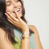 Pulsera ajustable de cordón de cuero color beige/camel con un dije central de corazón dorado de aspecto martillado. Presenta un cierre de bola dorada ajustable.