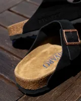 Sandalias planas de dos tiras ajustables, color negro, con hebillas metálicas color cobre. La plantilla es de corcho y látex, con forro de gamuza.