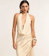 Vestido largo de satén color crema, con escote halter, espalda descubierta y abertura frontal. Presenta detalles de pedrería en el escote.
