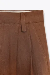 Pantalón marrón de tiro medio, confeccionado en mezcla de lino y viscosa. Presenta trabillas en la cintura, bolsillos laterales, pinzas y raya planchada en el delantero. Cierre frontal con cremallera, botón y ganchos metálicos.