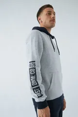 Buzo canguro gris con capucha ajustable y bolsillo delantero. Tiene puños y cintura acanalados en azul marino, y un parche con texto en el pecho.