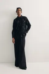 Camisa negra con textura, cuello de pico, botones delanteros, canesú con pliegues en la espalda, mangas largas con hombros caídos, puños abotonados y bajo redondeado.