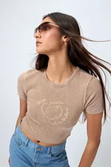 Remera color beige con cuello redondo y mangas cortas. Presenta un estampado con relieve de un corazón con un caballo en su interior y la frase "wild hearts can't be broken".
