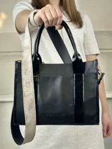 Bolso negro de cuero con detalles charolados, doble asa corta y correa larga de tela con el nombre de la marca.