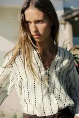 Blusa color crema con rayas verticales bordadas en negro. Tiene cuello redondo con escote en pico, mangas largas abullonadas y cierre frontal con botones.