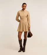 Vestido corto color beige, estilo chemise, confeccionado en tricolina. Tiene cuello inglés, mangas largas con puños abotonados, abotonadura frontal y falda con pliegues.