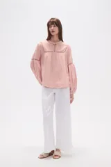 Blusa holgada de algodón color rosa pálido, con escote redondo y lazo ajustable en el centro. Presenta detalles de encaje calado alrededor del escote y en los puños de las mangas largas abullonadas.