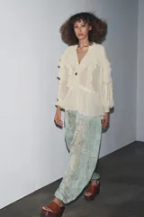 Blusa color crudo confeccionada en viscosa, con cuello redondo, escote en pico, mangas largas con puños elásticos, volantes frontales y detalles de encaje.