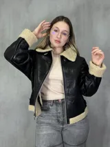 Campera negra de cuero sintético con interior y cuello de corderito color beige, cierre frontal con cremallera y hebillas ajustables en el cuello y la cintura.