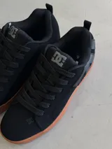 Championes DC Court Graffik color negro con suela de goma marrón. Presentan cordones gruesos negros, logo de DC en la lengüeta y en el lateral, y detalles de costuras.