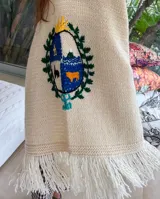 Ruana de lana merino color crudo, tejida y bordada a mano con el escudo de Uruguay en la espalda. Tiene calce holgado y flecos en el ruedo.