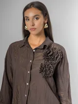 Camisa de corte clásico, color marrón oscuro con brillo sutil dorado, confeccionada en tela nacarada. Presenta apliques de flores tridimensionales fruncidas en el área del pecho.