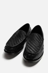 Mocasín de cuero negro con diseño trenzado en la parte superior, horma redonda y suela con ligero tacón.