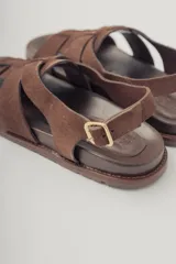 Sandalia plana de gamuza color marrón oscuro, con tiras cruzadas en la parte delantera y una tira fina ajustable con hebilla metálica en el tobillo. Posee plantilla de cuero y suela baja.