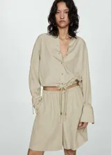 Camisa color beige de corte crop, confeccionada en mezcla de lino y lyocell. Tiene cuello camisero, cierre frontal con botones, manga larga con puños abotonados y lazo ajustable en la cintura.