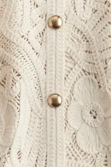 Saco corto color crudo, tejido al crochet con diseño floral. Tiene cuello redondo, mangas largas y cierre frontal con botones dorados.