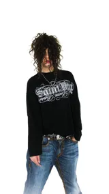 Remera negra oversize de manga larga, confeccionada en jersey de algodón grueso. Presenta cuello ancho y estampado frontal con el logo de la colaboración LUCCABOSSIO x Saint.
