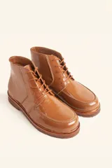 Bota de cuero vacuno con acabado charol, color marrón oscuro. Presenta un diseño de caña corta con cordones frontales y costuras decorativas en la puntera. La suela es de goma tipo crepe.