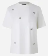 Blusa color crudo de algodón con cuello redondo y mangas cortas. Presenta bordados de lazos con perlas artificiales.