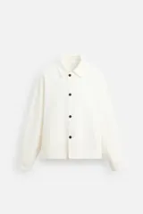 Sobrecamisa color crudo de corte boxy fit, confeccionada en tejido de algodón denim con estructura irregular. Tiene cuello solapa, manga larga con puños abotonados, bolsillos de vivo en la cadera y cierre frontal con botones.