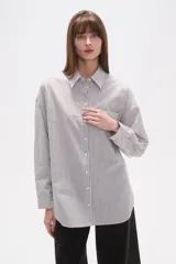 Camisa de algodón blanca de corte holgado, con cuello camisa y manga larga. Presenta un estampado de finas rayas verticales negras y un bolsillo frontal con rayas horizontales.