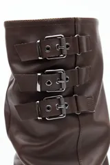 Bota de caña alta confeccionada en poliuretano con diseño de efecto fruncido y puntera redondeada. Presenta tres hebillas metálicas decorativas en el lateral superior de la caña y taco bajo.